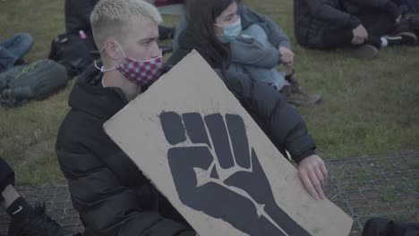 White-young-man-sitting-on-the-grass-holding-a-banner-with-a-black-fist-during-black-lives-matter-protest-in-Cambridge,-England