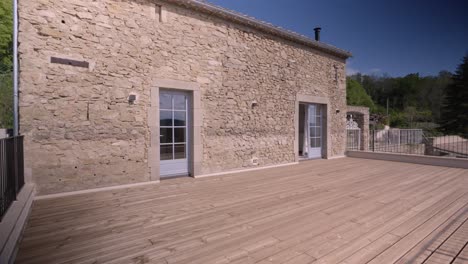 Große-Terrasse-Mit-Holzboden-Und-Zäunen,-Von-Einem-Haus-Mit-Steinmauern,-Zwei-Hellen-Französischen-Fenstern,-Blauer-Himmel-Und-Sonniges-Wetter,-Grün-Im-Hintergrund