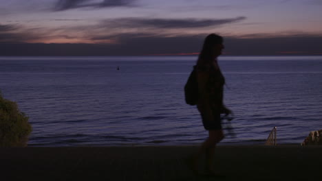 Das-Meer-Und-Der-Strand-Kurz-Nach-Sonnenuntergang-Mit-Vorbeigehenden-Menschen,-Die-Sich-Vor-Dem-Wunderschönen-Hellen-Himmel-Abheben