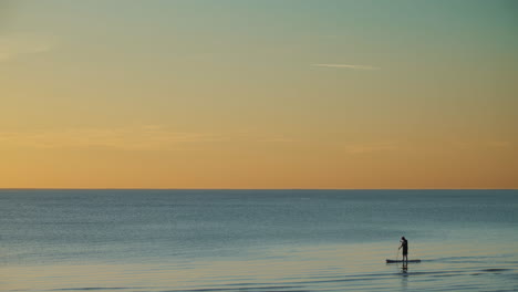 Person-stand-up-paddleboarding-at-sunset-on-calm-sea-waters,-summer-background