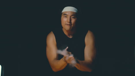 Asian-male-hyped-up-for-workout,-clapping-chalk-and-yelling-in-dark-gym,-4k-800fps-strong-athlete-wearing-headband-excited