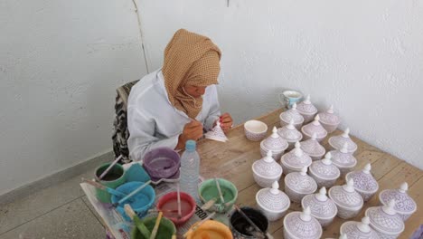 Eine-Arabische-Kunsthandwerkerin-Malt-In-Einer-Keramikwerkstatt-In-Fez-Detaillierte-Marokkanische-Muster-Auf-Tontöpfen-Von-Hand-Mit-Verschiedenen-Pinseln-Und-Farben