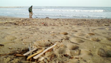 Man-walks-With-A-Metal-Detector-on-a-Sandy-Beach-On-The-Seashore-full-of-garbage-waste-After-a-Storm