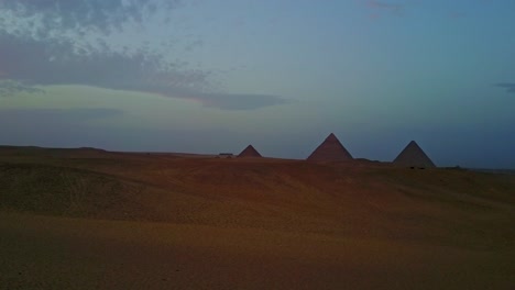 Toma-Aérea-Panorámica-A-Través-De-Las-Arenas-Del-Desierto-En-Egipto-Con-Las-Pirámides-Al-Fondo-Durante-El-Anochecer.