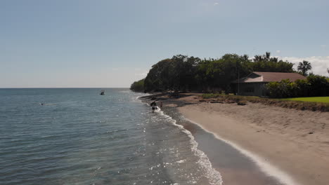 A-drone-flies-slowly-over-the-gentle-waves-of-Olowalu-Beach-as-a-father-and-two-boys-walk-on-the-beach-in-Maui,-Hawaii