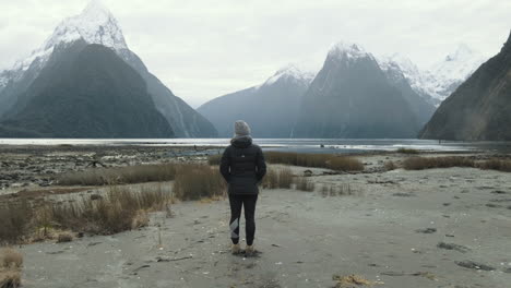 Girl-standing-alone-in-front-of-towering-snow-capped-mountains-and-fiords-in-New-Zealand's-Milford-Sound