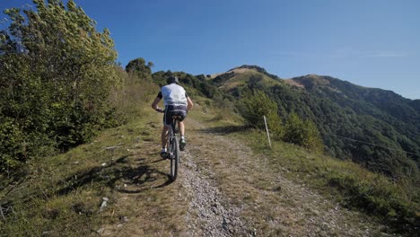 Male-mountain-biker-cycling-uphill-on-gravel-road,-looking-towards-mountains-on-sunny-summer-day,-slow-motion-wide-shot