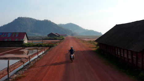 Drone-follows-biker-riding-on-dusty-dirt-road-between-farms-and-salt-plantations