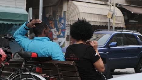 African-couple-sitting-on-a-bench-in-an-urban-street-area,-talking-while-playing-with-their-hair