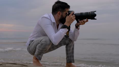 Un-Fotógrafo-Que-Toma-Fotografías-Agachado-En-Una-Playa-Con-Los-Pies-En-El-Agua