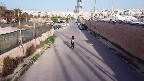 Active-track-drone-shot-of-a-young,-athletic,-caucasian-woman-inline-skating-on-the-streets-from-the-back-in-the-middle-of-parking-cars,-waving-her-arms-up-as-she-is-gliding-through-the-windy-breeze