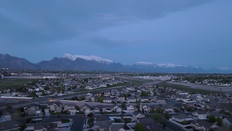 Crepúsculo-Sobre-Una-Expansión-Suburbana-En-Un-Valle-Debajo-De-Las-Montañas-Cubiertas-De-Nieve:-Revelación-Aérea-De-Retroceso