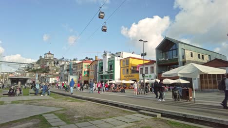 Crowded-street-in-Gaia,-Porto,-with-colorful-buildings-and-cable-cars-above,-people-walking,-sunny-day