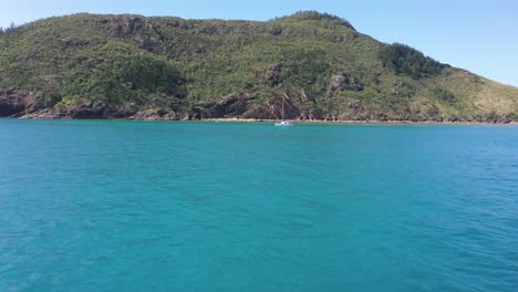 Low-aerial-over-blue-sea-to-white-catamaran-sailboat-moored-off-island