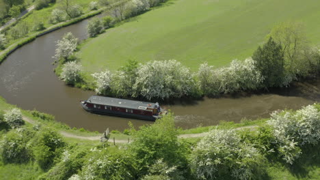 An-aerial-view-of-a-canal-boat-navigating-a-canal-corner,-surrounded-by-Yorkshire-countryside-on-a-sunny-spring-day