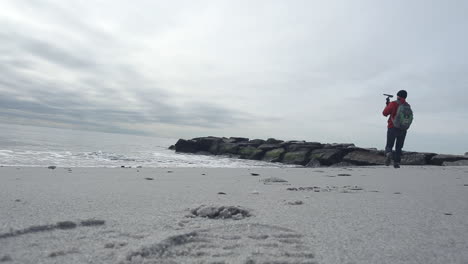 Male-photographer,-videographer-filming-landscape-scenery-of-the-ocean-on-a-cloudy-day-as-he-walks-on-the-beach-with-shoes-leaving-footsteps-on-the-sand