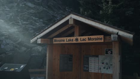 Small-Wooden-Structure-With-Moraine-Lake-Signage-At-The-Banff-National-Park-In-Alberta,-Canada-Revealed-Behind-Bokeh-Effect-On-A-Bright-Sunny-Day---Panning-Shot