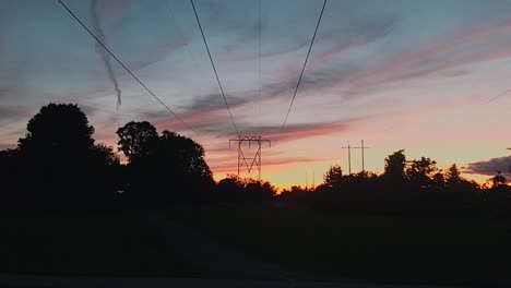 Time-lapse-of-the-sunset-over-a-trail-and-power-lines-with-trees-on-either-side-with-a-road-and-sidewalk-filled-with-cars-and-people-in-a-town-just-outside-of-Ottawa,-Ontario