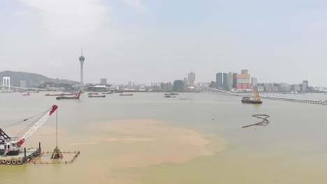 Ein-Hoher-Drohnen-Truck-Schoss-An-Einem-Greifbagger-Und-Einem-Hopperkahn-Vorbei,-Im-Hintergrund-Die-Skyline-Der-Stadt-Macau