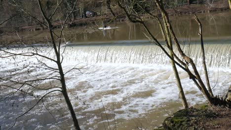 Person,-Die-Im-Wald-Auf-Einem-Ruhigen,-Seichten,-Braunen-Fluss-Mit-Wasserfall-Kajak-Fährt