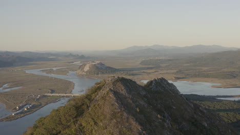 Pull-back-shot-of-summit-of-a-pyramid-shaped-mountain-with-reveal-of-a-river-estuary-and-mountain-ridge-in-the-background,-on-the-sunset