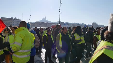 Demonstranten-In-Gelben-Jacken-Machen-Sich-Auf-Den-Weg-Zu-Einem-Roten-Krankenwagen-In-Marseille,-Mit-Notre-Dame-De-La-Garde-Im-Hintergrund-Unter-Einem-Perfekten-Blauen-Himmel-An-Einem-Sonnigen-Tag