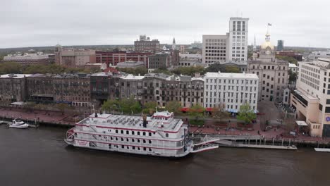 Aerial-low-panning-shot-of-a-riverboat-cruise-docked-on-the-Savannah-River-in-downtown-Savannah,-Georgia