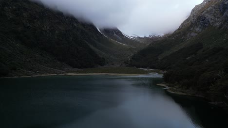 Vuelo-Vertical-De-Drones-Captura-Un-Lago-En-Medio-De-Montañas-Cubiertas-Por-Nubes