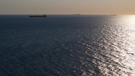 Tanker-container-ships-line-up-on-horizon,-silhouettes-of-boats-out-to-distance-as-drone-rises