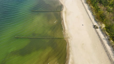 Vista-Aérea-De-Una-Playa-Serena,-Una-Clara-Demarcación-Entre-La-Costa-Arenosa-Y-Las-Aguas-Poco-Profundas,-Con-Dos-Embarcaderos-Que-Se-Extienden-Hacia-El-Mar,-Resaltando-El-Entorno-Natural-Tranquilo-Y-Tranquilo.