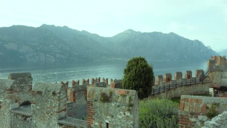 Impresionante-Vista-Del-Lago-De-Garda-Desde-Malcesine,-Que-Presenta-El-Efecto-De-Paralaje,-Con-Un-Telón-De-Fondo-Del-Majestuoso-Castillo,-Montañas-Rocosas-Y-árboles-Frondosos,-Que-Muestra-La-Rica-Cultura-Cultural.