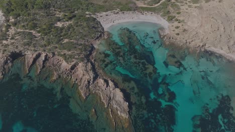 Aerial-panoramic-natural-blue-rocky-cliff-bay-forested-Cala-Sa-Torreta-Beach-Sea-in-Menorca-Spain,-travel-destination