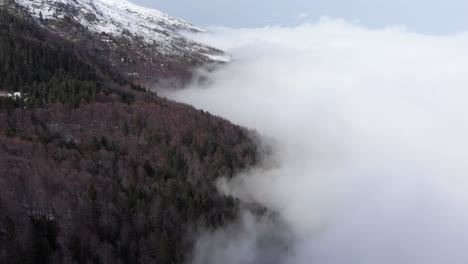 Drone-view-of-beautiful-mountainside-forest-covered-in-floating-clouds-snow-covered-mountain-peak-at-the-distance-winter-day-Kaimaktsalan-Greece