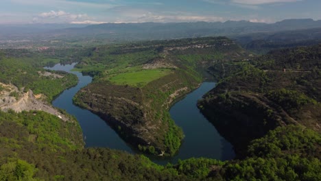 A-winding-river-in-salvassola-vic-near-barcelona,-lush-greenery-and-mountainous-backdrop,-aerial-view