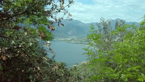 View-of-Lake-Garda-through-lush-vegetation-from-a-mountain-hiking-trail,-featuring-trees,-sky,-clouds,-and-water