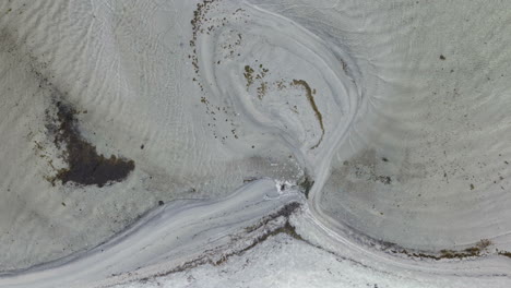 Vista-Aérea-De-Arriba-Hacia-Abajo-De-Una-Playa,-Donde-El-Agua-Fluye-A-Través-De-La-Arena-Creando-Hermosos-Patrones-Arremolinados.