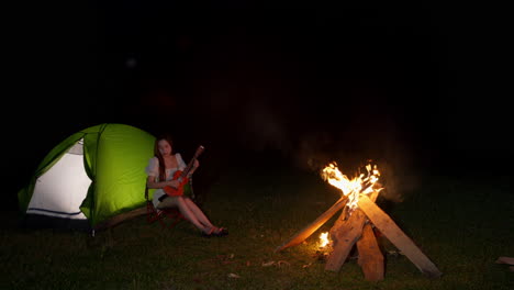 young-female-artist-playing-guitar-ukulele-while-camping-outdoor-in-front-of-bonfire-playing-a-song-under-the-moonlight