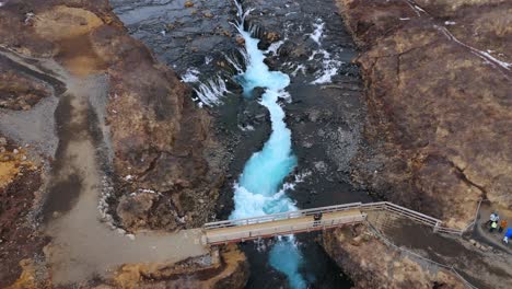 Bruarfoss-waterfall-in-iceland-with-vibrant-blue-water-and-wooden-bridge,-aerial-view