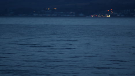 Calm-water-surface-with-ripples-from-current-and-shimmering-town-lights-in-background