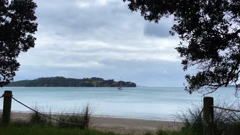 Blick-Vom-Grasbewachsenen,-Sandigen-Ufer-Auf-Ein-Segelboot,-Das-In-Der-Bucht-Ablegt,-Neuseeland