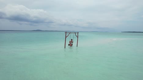 pretty-young-woman-relaxing-on-a-swing-in-turquoise-blue-water-with-sand-beach-at-Leebong-Island-in-Belitung-Indonesia,-aerial