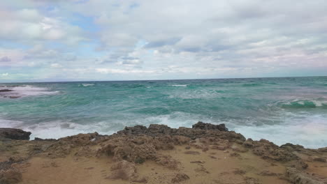 Las-Olas-Ruedan-Suavemente-Sobre-Una-Playa-Rocosa-Bajo-Un-Cielo-Parcialmente-Nublado,-Capturando-El-Sereno-Movimiento-Del-Mar-En-Cámara-Lenta.