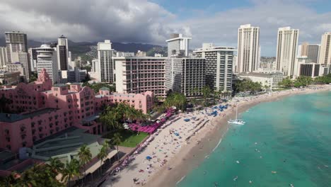 Aerial-shot-along-Honolulu's-Waikiki-Beach,-Hawaii,-pristine-turquoise-waters-with-resorts,-tourists,-swimmers-and-surfers