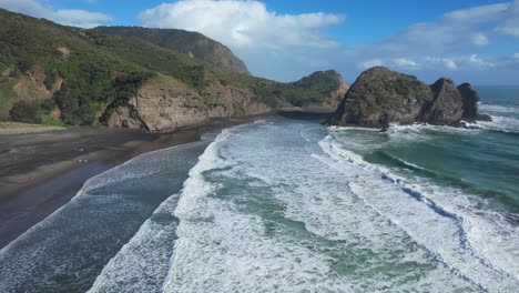 Waves-Over-The-Black-Sand-Beach-Of-Piha-With-Tasman-Lookout-And-Taitomo-Rock-In-Auckland-Region,-North-Island,-New-Zealand