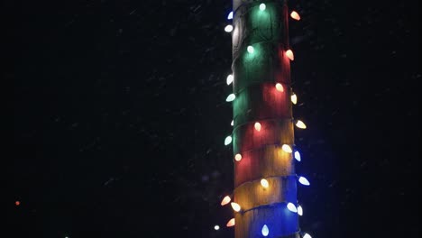 Winter-snowfall-at-night-snowing-on-Christmas-lights-on-city-street-in-street-light