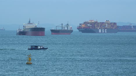 Buques-Portacontenedores-Anclados-En-El-Estrecho-De-Singapur-Mientras-Barcos-De-La-Autoridad-Gubernamental-Navegan,-Inspeccionando-Mercancías-Aduaneras-Y-Documentando-Su-Entrada-Al-País.