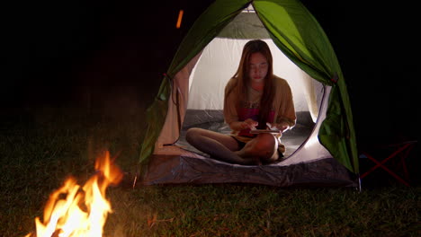 Mujer-Asiática-Acampando-Al-Aire-Libre-Charlando-Dentro-De-Su-Tienda-Con-Un-Moderno-Teléfono-Inteligente-Conectado-A-5g-Frente-A-La-Hoguera