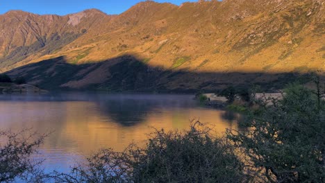 Shadow-from-a-mountain-cast-across-a-lake-from-early-morning-sun,-with-fog-and-a-fisherman