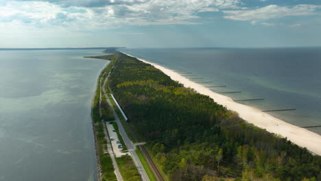 A-breathtaking-aerial-view-along-the-Hel-Peninsula-in-Kuźnica,-showing-a-verdant-forest-strip-sandwiched-between-a-serene-bay-and-the-Baltic-Sea,-with-a-visible-railway-and-roadway-running-parallel