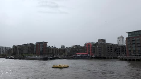 Sailing-Past-Butlers-Wharf-And-Apartments-On-South-Bank-Of-River-Thames-On-Overcast-Cloudy-Day